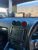 A circular black car dashboard accessory with a red illuminated ring, mounted on the center console above the radio, shown inside a vehicle.
