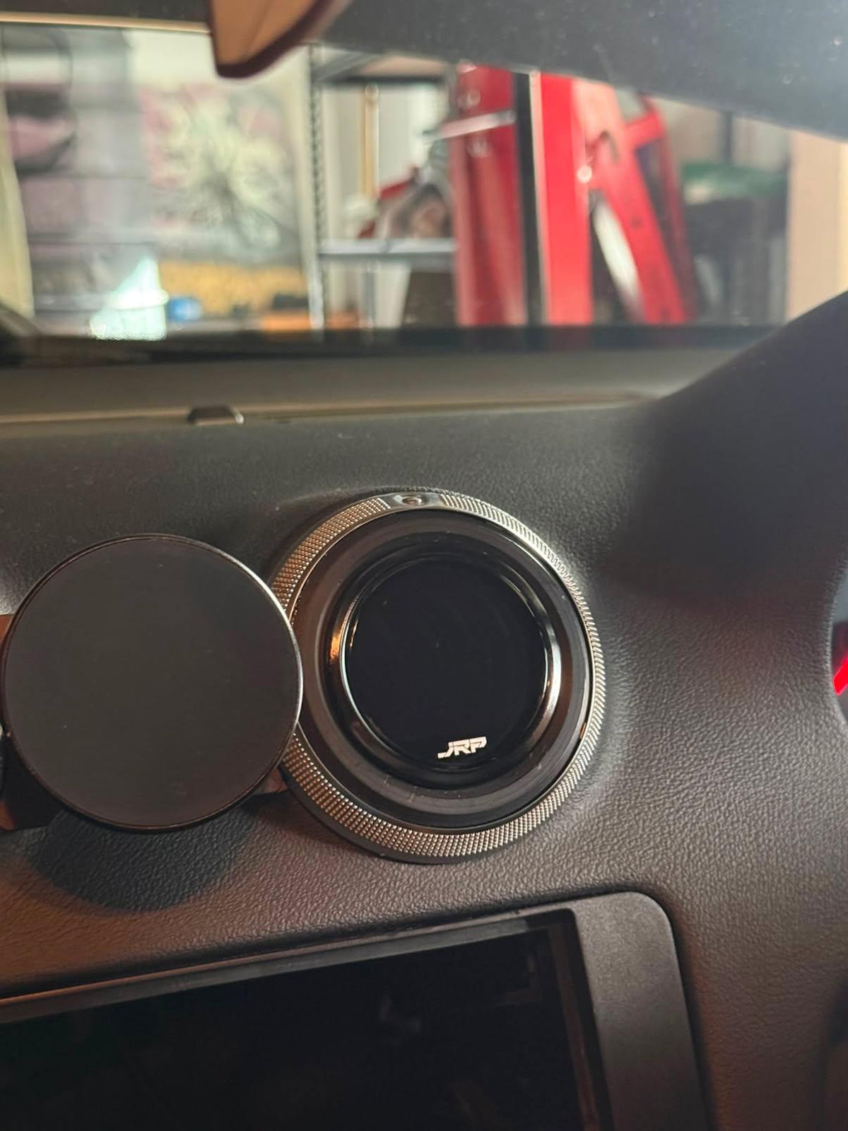 A close-up of a circular car air vent cover installed on a dark gray dashboard, with a matching round black cover on the left. The vent has a textured silver outer ring and a small white 'JYP' logo at the bottom.